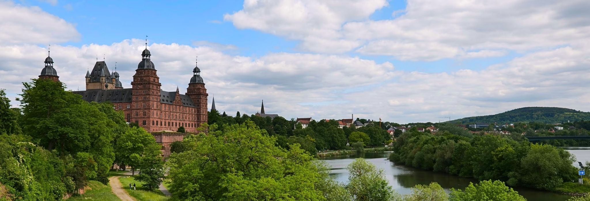 Weitläufige Ansicht von Schloss Johannisburg und dem grünen Mainufer unter einem bewölkten Himmel. Diese regionale Kulisse unterstreicht unsere Verbundenheit bei der Planung und Montage für Ihre PV Anlage Aschaffenburg.