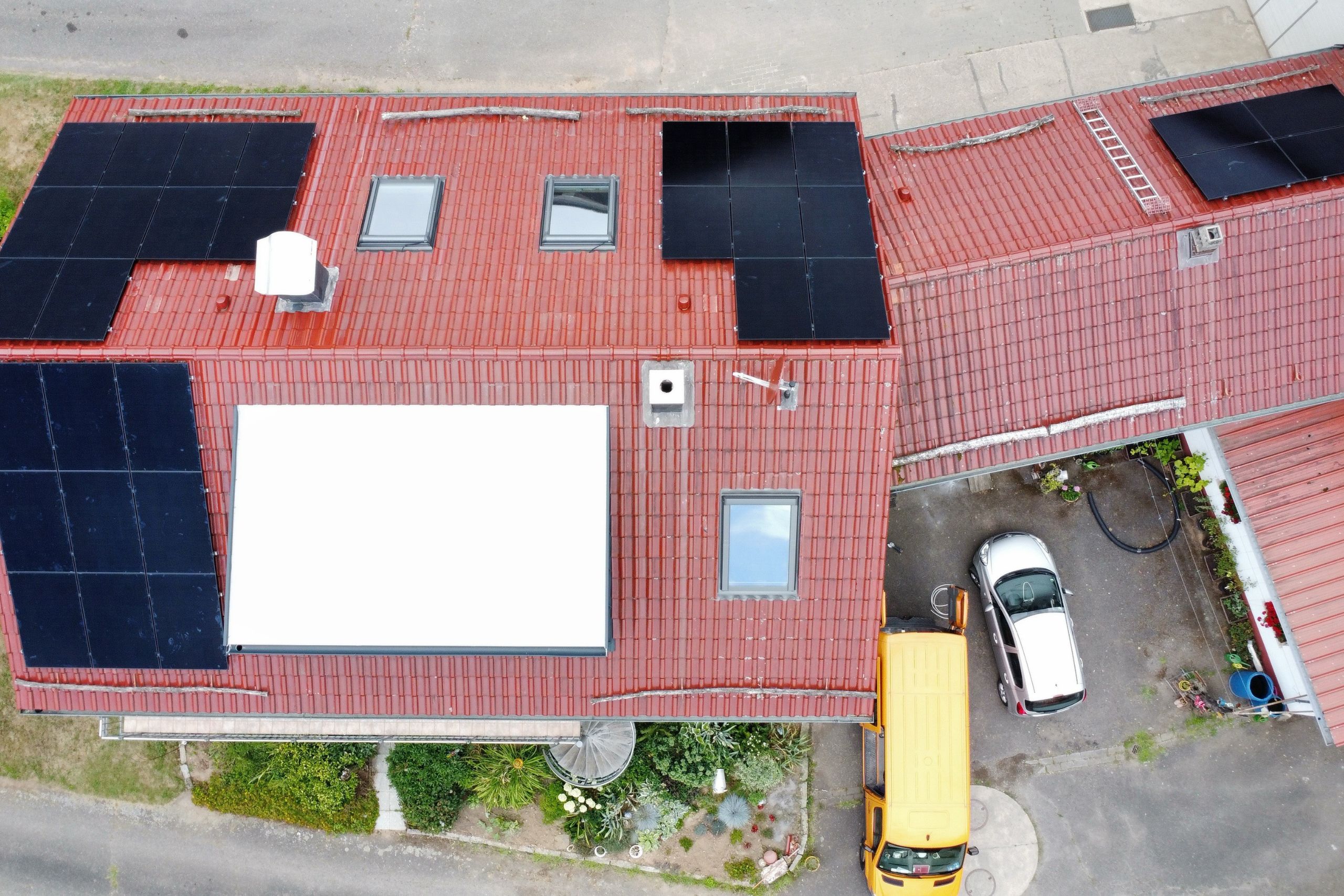 PV Anlage Aschaffenburg Wohnhaus Komplettdach Luftaufnahme eines Wohnhauses mit einem Satteldach, das fast vollständig von einer großflächigen, modernen Photovoltaikanlage bedeckt ist. Ein Referenzprojekt für effiziente PV Anlagen in Aschaffenburg, umgeben von grünen Gärten unter blauem Himmel.