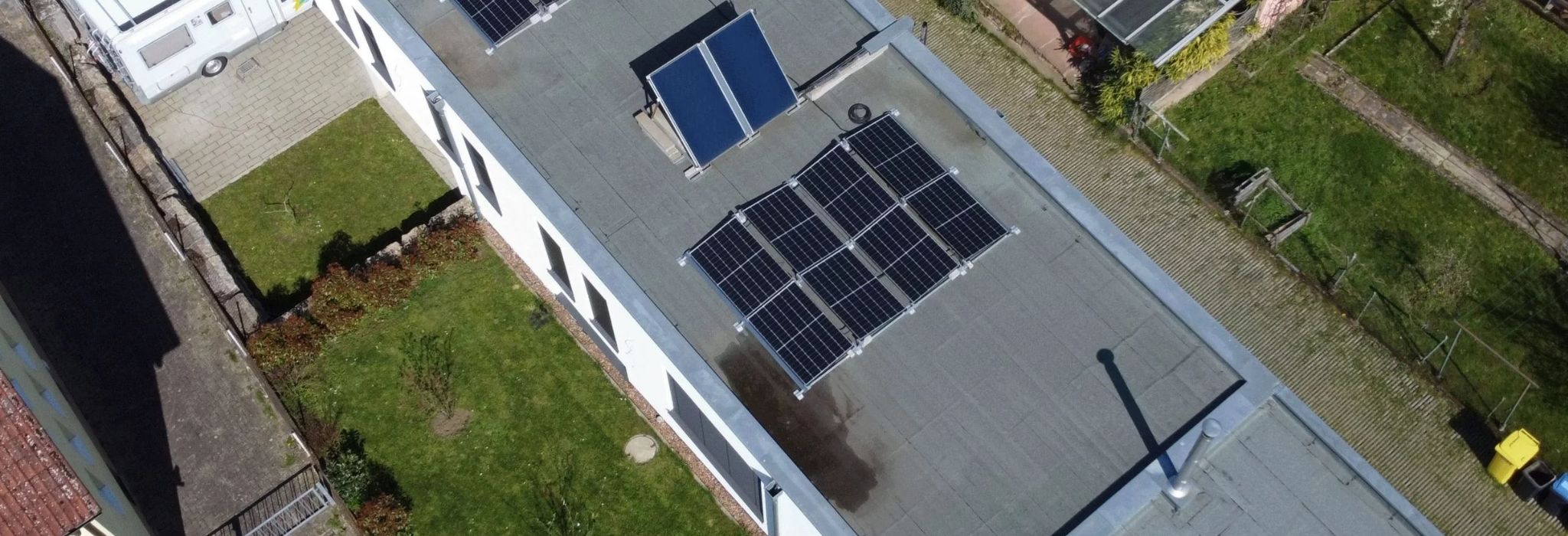 Detaillierte Luftaufnahme eines modernen Wohnhauses mit Flachdach. Aufgeständerte Photovoltaik-Module und Solarthermie-Kollektoren sind fachgerecht und optimal ausgerichtet installiert. Im Hintergrund sind ein Garten und ein Wohnmobil zu sehen. Dieses Projekt ist ein starkes Beispiel unserer Referenzen für eine PV Anlage in Miltenberg.