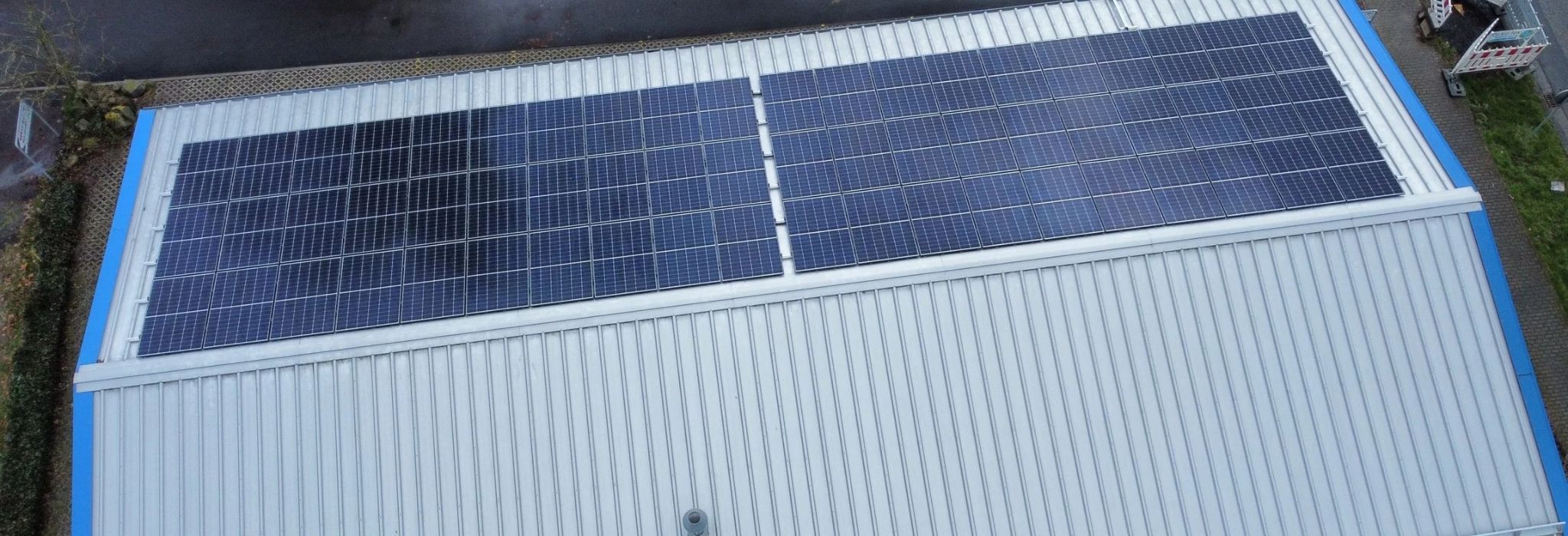 Detaillierte Drohnenaufnahme eines großen Flachdachgebäudes, das von Gerüsten umgeben ist. Eine umfangreiche Photovoltaikanlage ist teilweise montiert. Die Aufnahme zeigt die teils fertigen Modulfelder neben Bereichen mit Lücken und sichtbaren Unterkonstruktionen sowie Baumaterialien, die die laufende professionelle Installation dieser PV Anlage Rhein-Main dokumentieren.